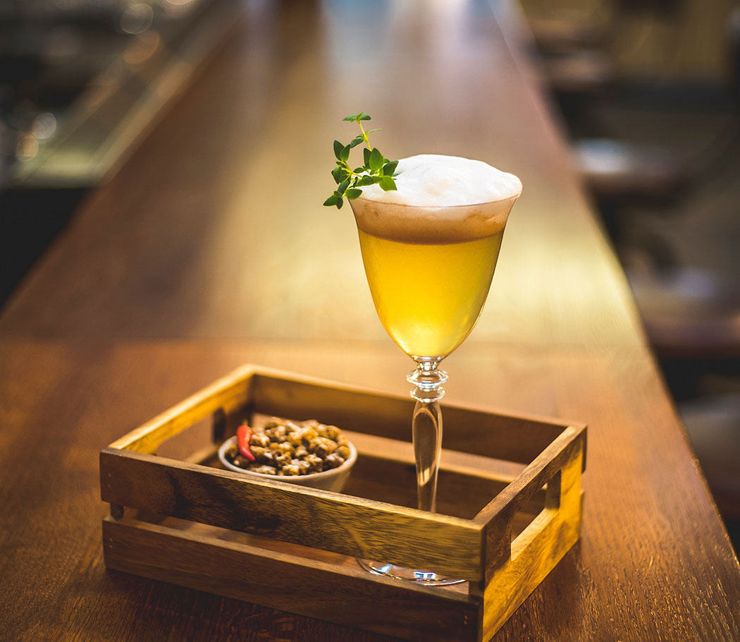 A filled beer glass sits inside a toy sized wooden crate, with some bar fare placed beside. This food and drink combination rests on the main bar.